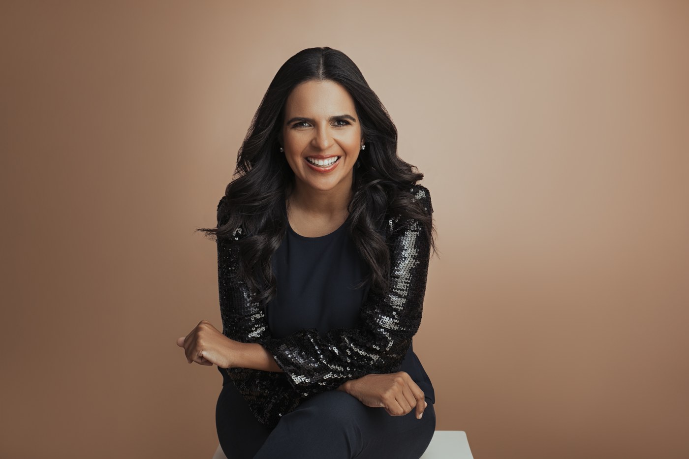 Janis Santaella sentada y sonriente, vestida de negro sobre un fondo beige